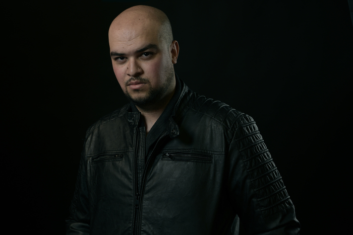 Photograph of Kevin Van Whye, a young, mixed-race man with a bald head and neatly trimmed beard. He is wearing a black jacket and stands before a black background.