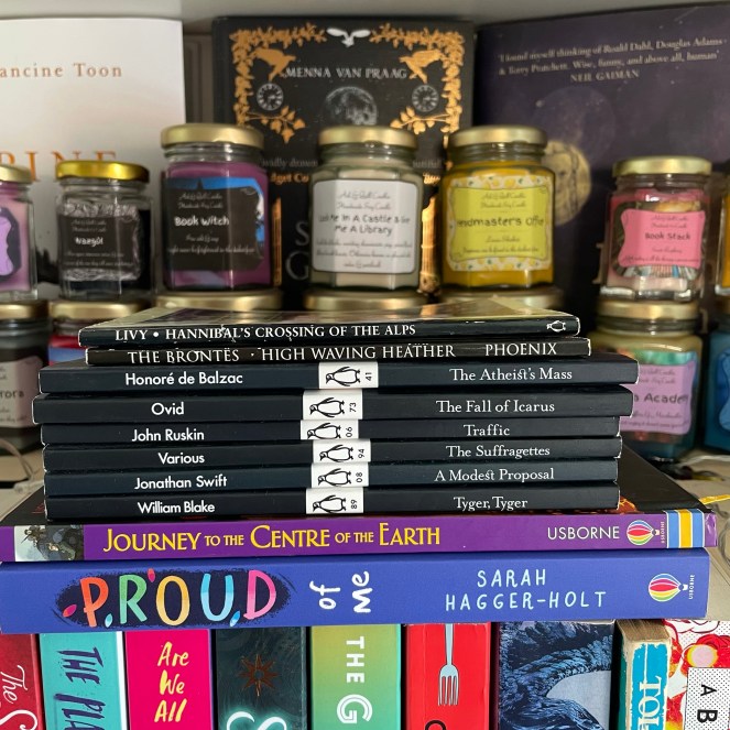 Image of a stack of books on a bookshelf with book-themed scented candles in the background.

The stack contains the following titles:
Hannibal's Crossing Of The Alps - Livy
High Waving Heather - The Brontes
The Atheist's Mass - Honore de Balzac
The Fall Of Icarus - Ovid
Traffic - John Ruskin
The Suffragettes - Various Authors
A Modest Proposal - Jonathan Swift
Tyger, Tyger - William Blake
Journey To The Centre Of The Earth
Proud Of Me - Sarah Hagger-Holt