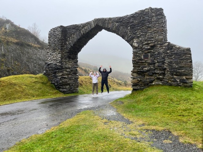 Image shows Carole and Caius in rain coats, jumping in the air beneath a stone arch on a rainy and foggy day in Wales.