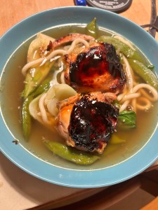 A blue bowl sits on the table. It is filled with a home-made clear broth, thick udon noodles, sugar snap peas, bok choi, and teriyaki-glazed chicken thighs.
