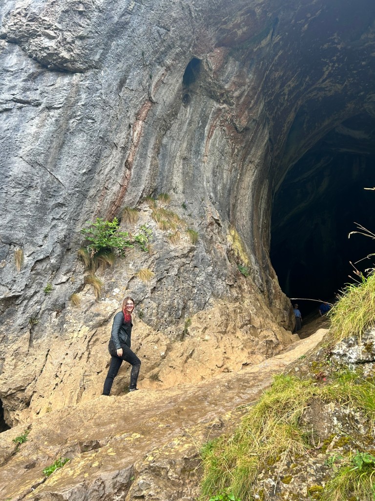 Tori stands at the entrance to Thor's Cave. She is wearing dark jeans and a leather jacket, and is smiling at the camera.