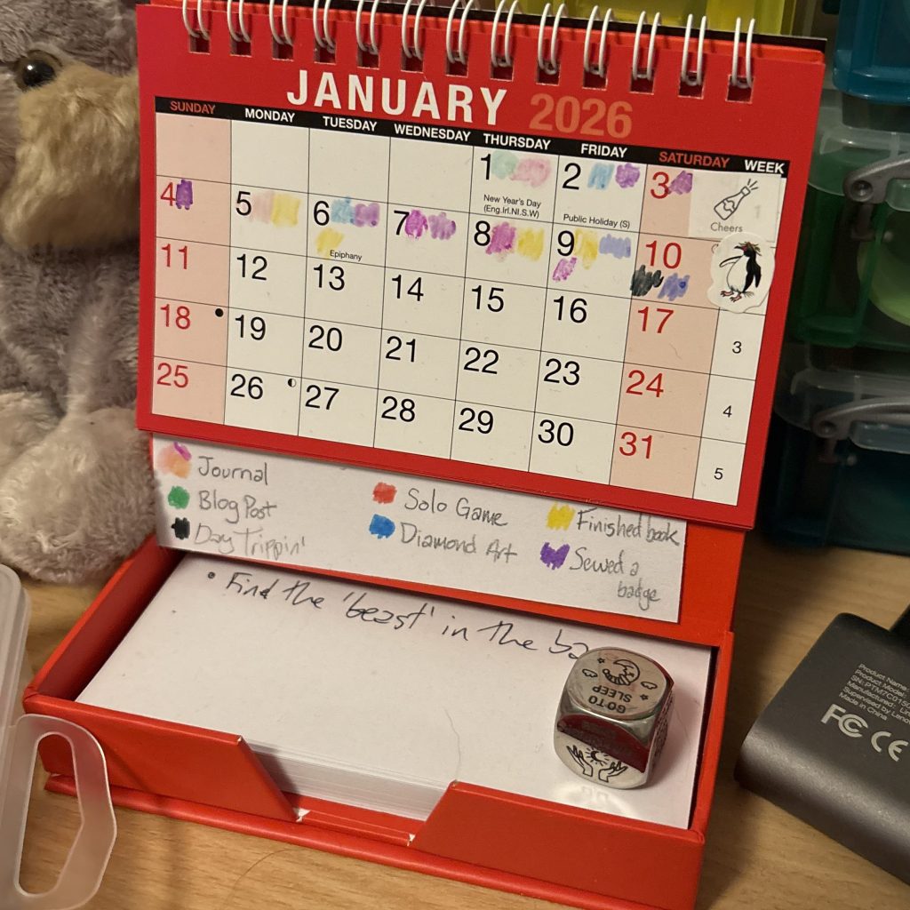 A small red desk calendar with the short form version of the aforementioned list colour-coded underneath it. The first 10 days have colour splodges on to show what I have done.
The base of the calendar has notepaper in which says 'Find the beast in the bay' and a chunky metal 'decision dice' sat in it.